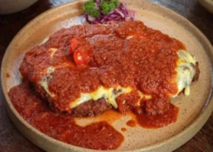 Bife à parmegiana empanado e coberto com queijo derretido e molho de tomate encorpado, servido num prato cerâmico rustico com guarnição de repolho roxo e salsinha.