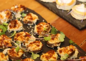 Cogumelos recheados e gratinados com queijo, salpicados com sementes de sésamo e brotos verdes, servidos numa tábua de ardósia escura.