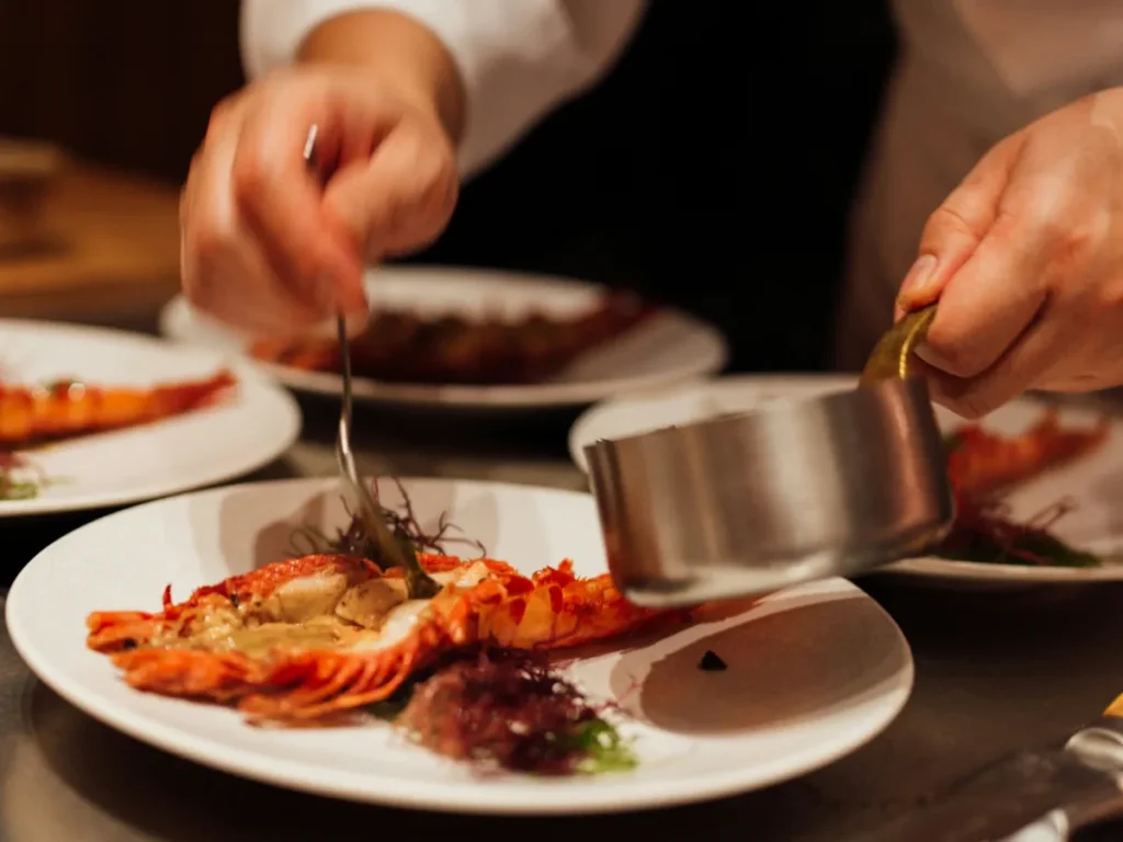 Fotografia em plano detalhe das mãos de um chef finalizando um prato de lagosta grelhada, derramando molho com uma pequena panela de inox em um restaurante.