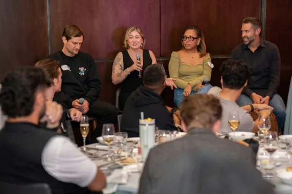 Mesa de debate em evento corporativo com quatro palestrantes sentados ao fundo e público em primeiro plano assistindo a uma palestra durante um jantar.