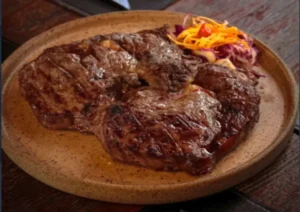 Corte alto de entrecôte grelhado na grelha, com marcas bem definidas de tostagem, servido num prato cerâmico rústico acompanhado de salada de cenoura ralada e repolho roxo.