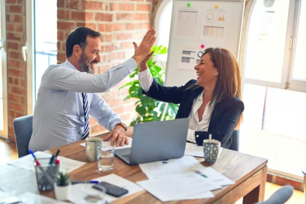 Homem e mulher em trajes executivos comemorando com um "high five" em uma mesa de reunião com notebook e documentos, simbolizando parceria de sucesso.