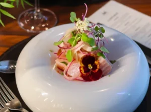 Ceviche de peixe branco com cebola roxa, brotos verdes e flor comestível num prato fundo de cerâmica branca.
