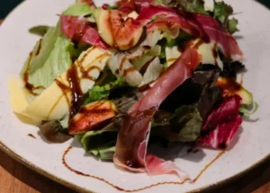 Salada de rúcula e folhas mistas com presunto cru, figos frescos em fatias, queijo em lascas e cobertura de vinagrete balsâmico cremoso num prato de cerâmica rústico.