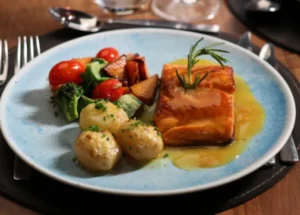 Lombo de salmão grelhado com molho cítrico e alecrim, acompanhado de batatas salteadas, brócolos e tomates-cereja num prato azul-claro.
