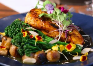 Peito de frango grelhado com molho de mostarda em grãos, acompanhado de brócolis, cogumelos, amêndoas laminadas e flores comestíveis num prato azul.