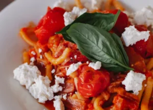 Prato de massa artesanal ao molho de tomate vermelho, finalizado com queijo ricota esfarelado, tomates-cereja assados e folhas frescas de manjericão no topo.