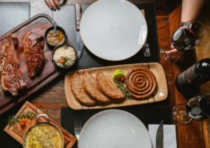 Mesa de refeição vista de cima com cortes de carne grelhada numa tábua de madeira, linguiça em espiral com fatias de pão, acompanhamentos e taças de vinho tinto.